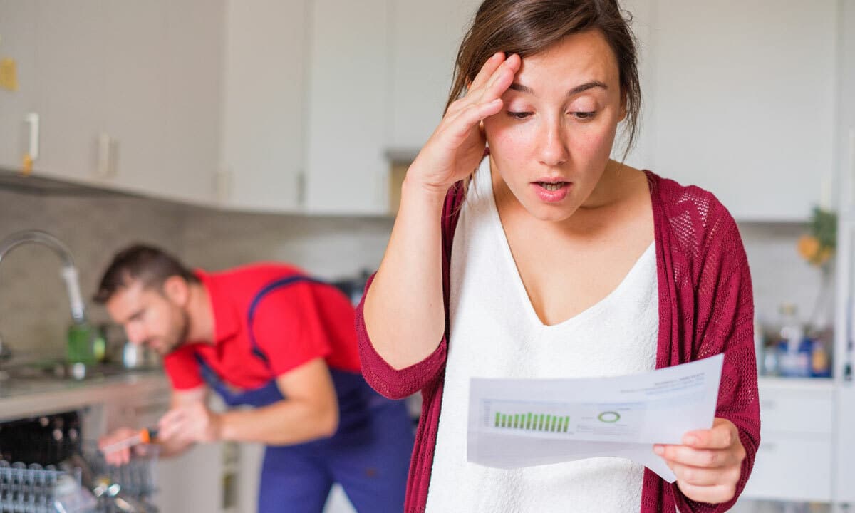 A woman shocked at the bill while the plumber works in the background